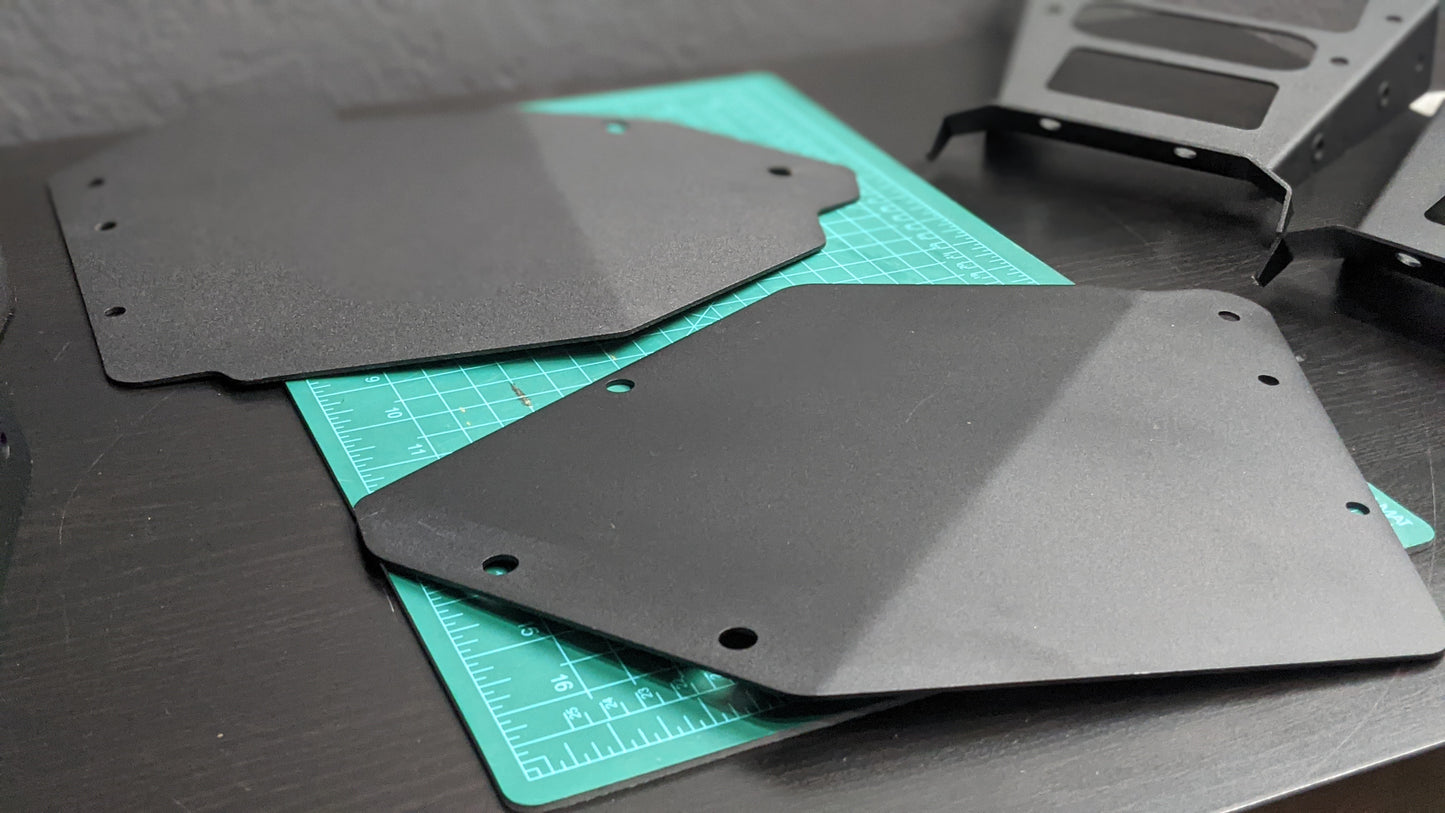 Two StayFlush Clean Slate Console – Universal Blank Build Platforms from StayFlush Motorsports with cut corners and holes rest on a green cutting mat on a dark desk, with additional metal components in the background.