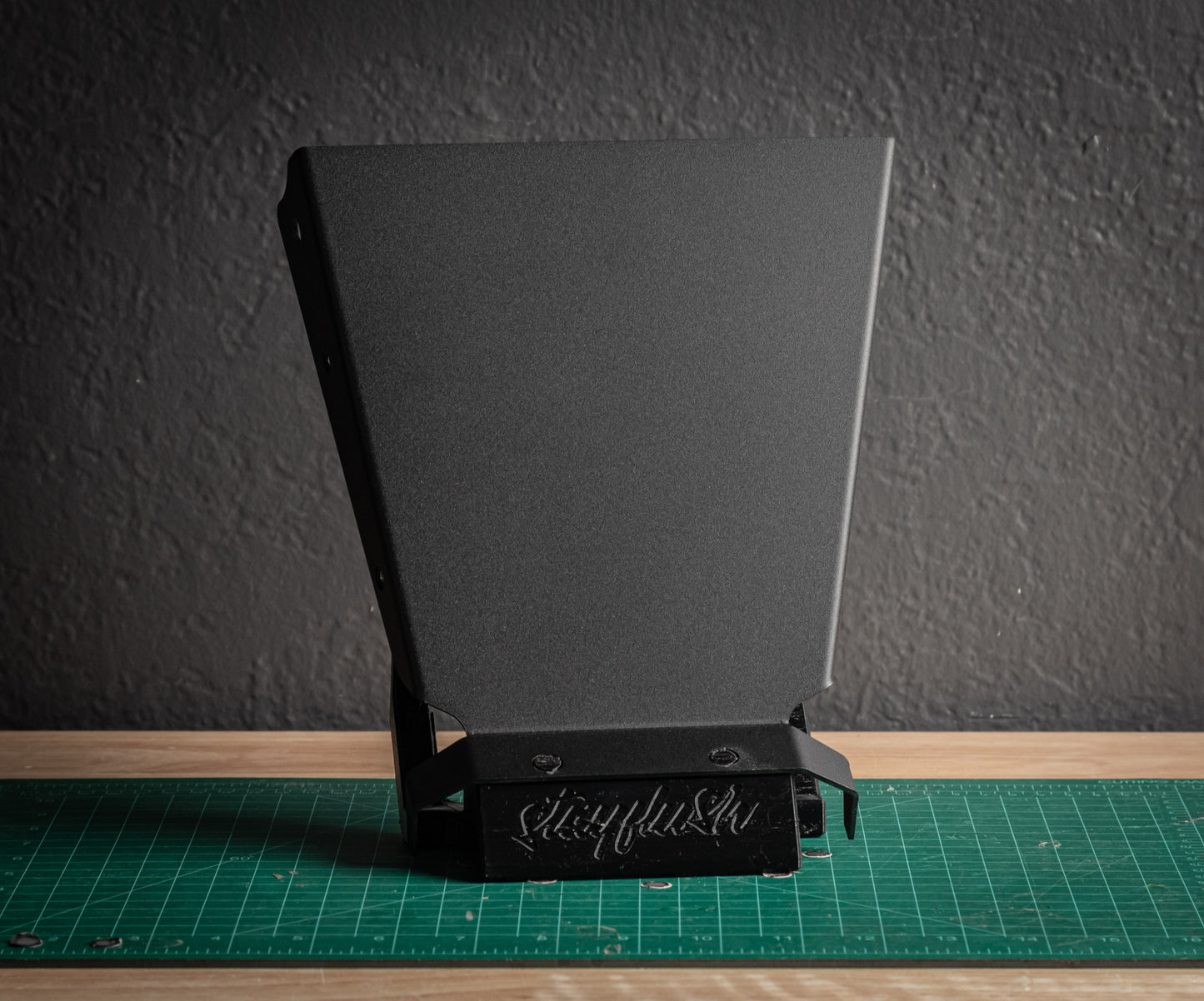 A black StayFlush Clean Slate Console – Universal Blank Build Platform by StayFlush Motorsports sits on a green cutting mat against a gray wall, with "Sikkyfab" engraved on its base.