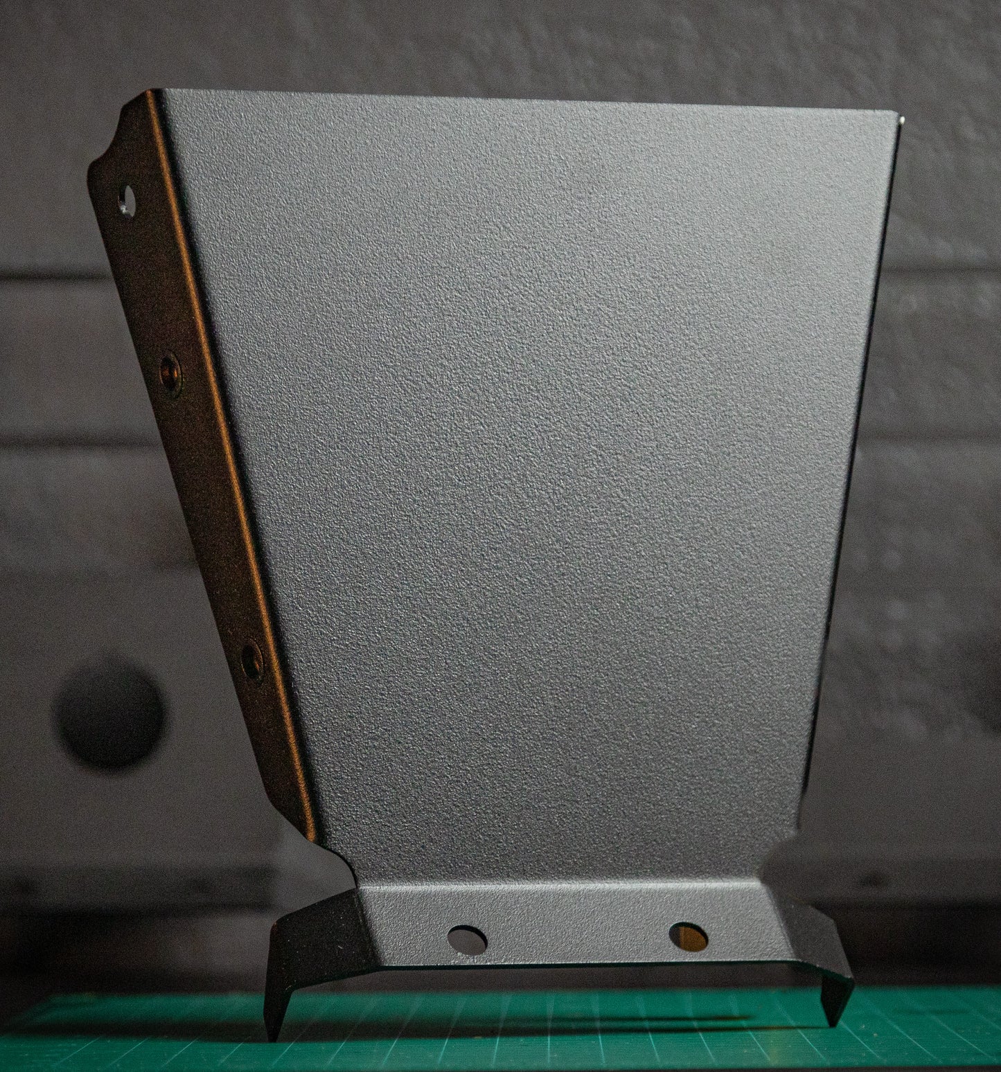 The StayFlush Clean Slate Console – Universal Blank Build Platform by StayFlush Motorsports stands upright on a green cutting mat, showing its textured metal surface, angled edges, and two holes against a blurred gray background.