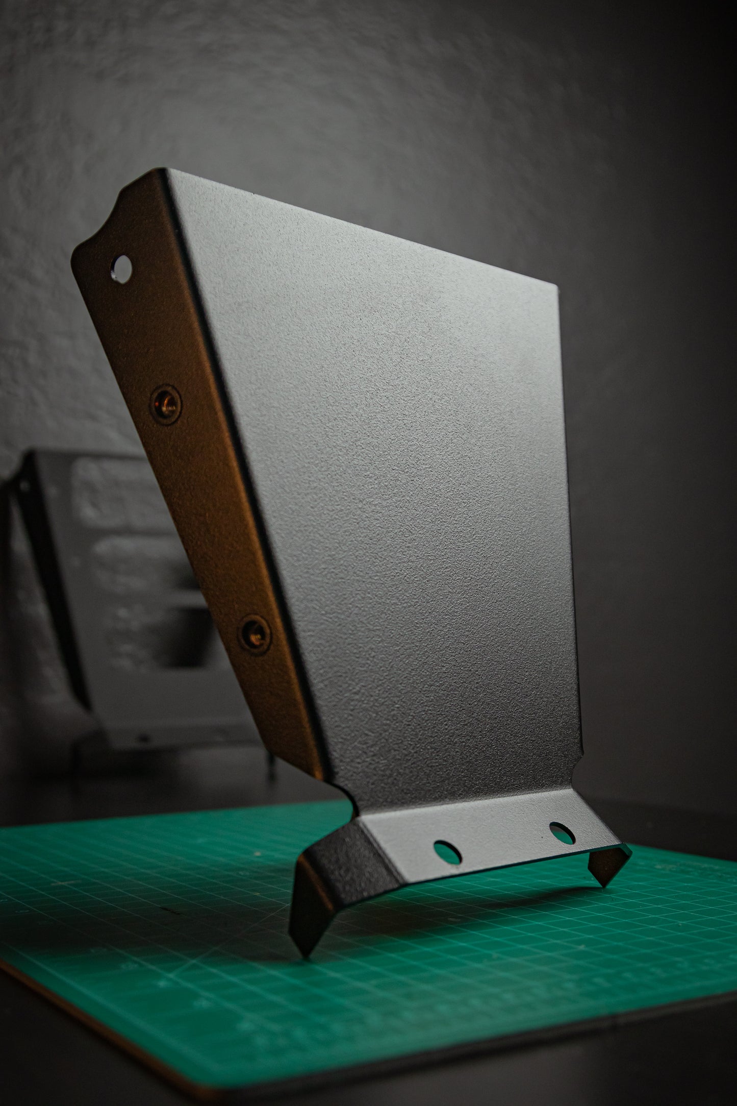 The StayFlush Clean Slate Console – Universal Blank Build Platform by StayFlush Motorsports is shown upright on a green cutting mat, with its angled black metal edges and mounting holes, while another console appears blurred in the background.