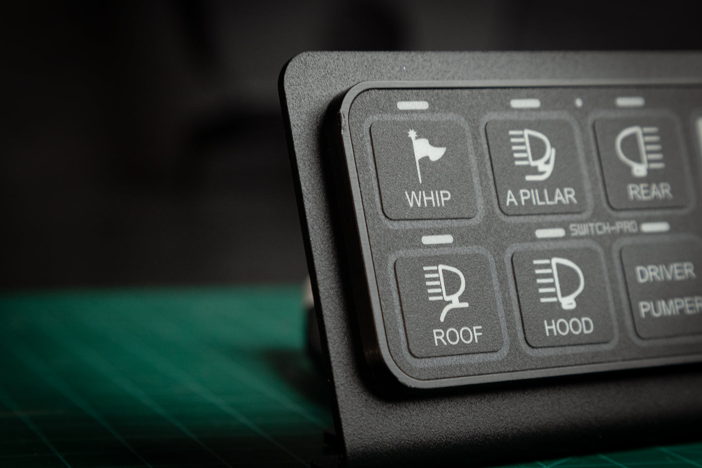 Close-up of the Switch-Pros 9100 Sidekick Accessory Mount by StayFlush Motorsports, showing labeled buttons—WHIP, A PILLAR, REAR, ROOF, and HOOD with icons—on a green cutting mat.
