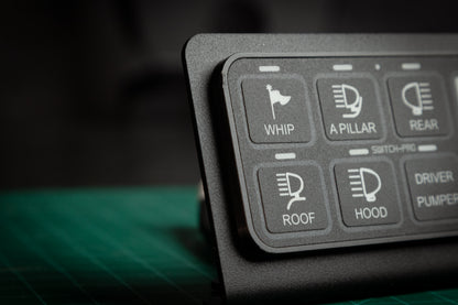 Close-up of the Switch-Pros 9100 Sidekick Accessory Mount by StayFlush Motorsports, showing labeled buttons—WHIP, A PILLAR, REAR, ROOF, and HOOD with icons—on a green cutting mat.
