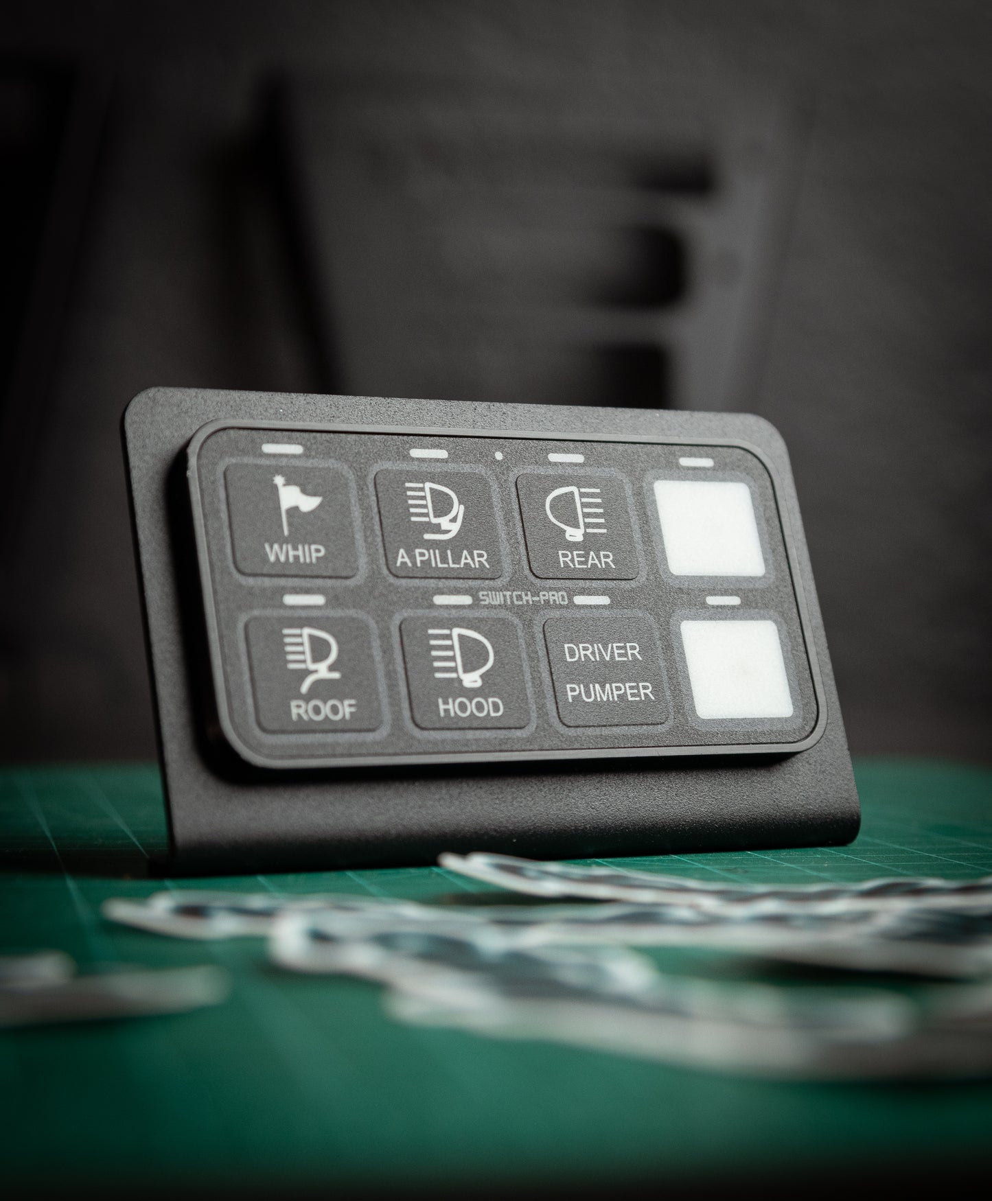 The Switch-Pros 9100 Sidekick Accessory Mount by StayFlush Motorsports is a black, rectangular switch panel with labeled buttons displayed on a green surface against a dark, blurred background.