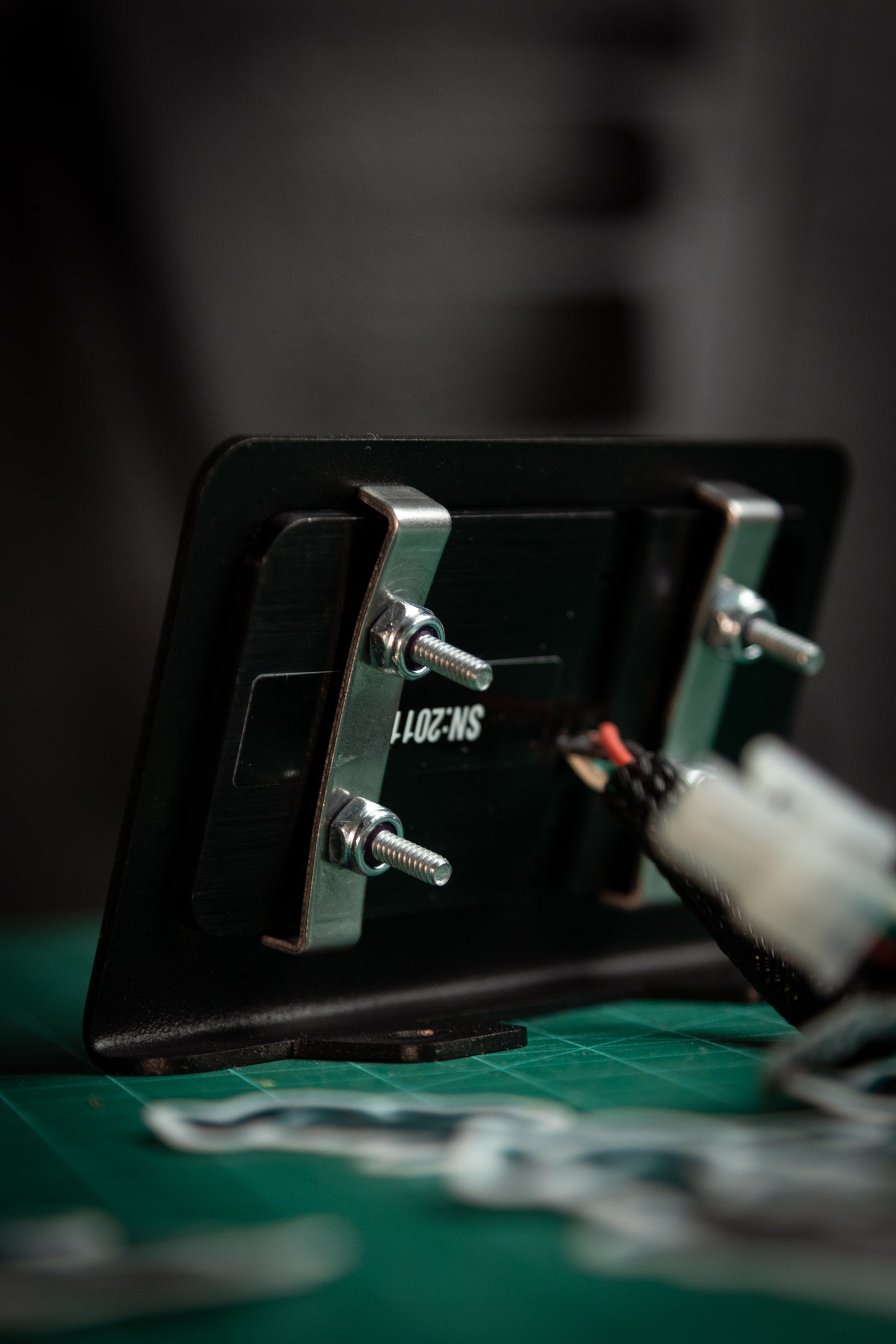 A close-up of the Switch-Pros 9100 Sidekick Accessory Mount by StayFlush Motorsports, a black rectangular metal plate labeled SN:2011, secured with bolts and partially hidden by wires on a green surface.