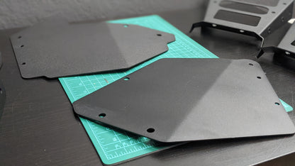 Two StayFlush Clean Slate Console – Universal Blank Build Platforms from StayFlush Motorsports with cut corners and holes rest on a green cutting mat on a dark desk, with additional metal components in the background.