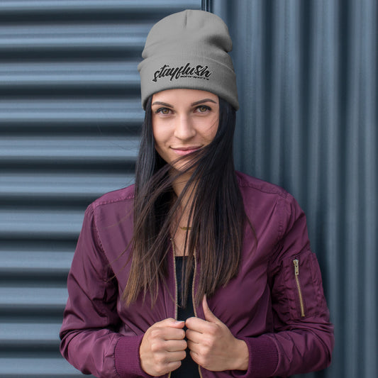 A woman in a maroon jacket and gray StayFlush Beanie stands in front of a corrugated metal wall, looking at the camera with a slight smile and some hair partially covering her face.