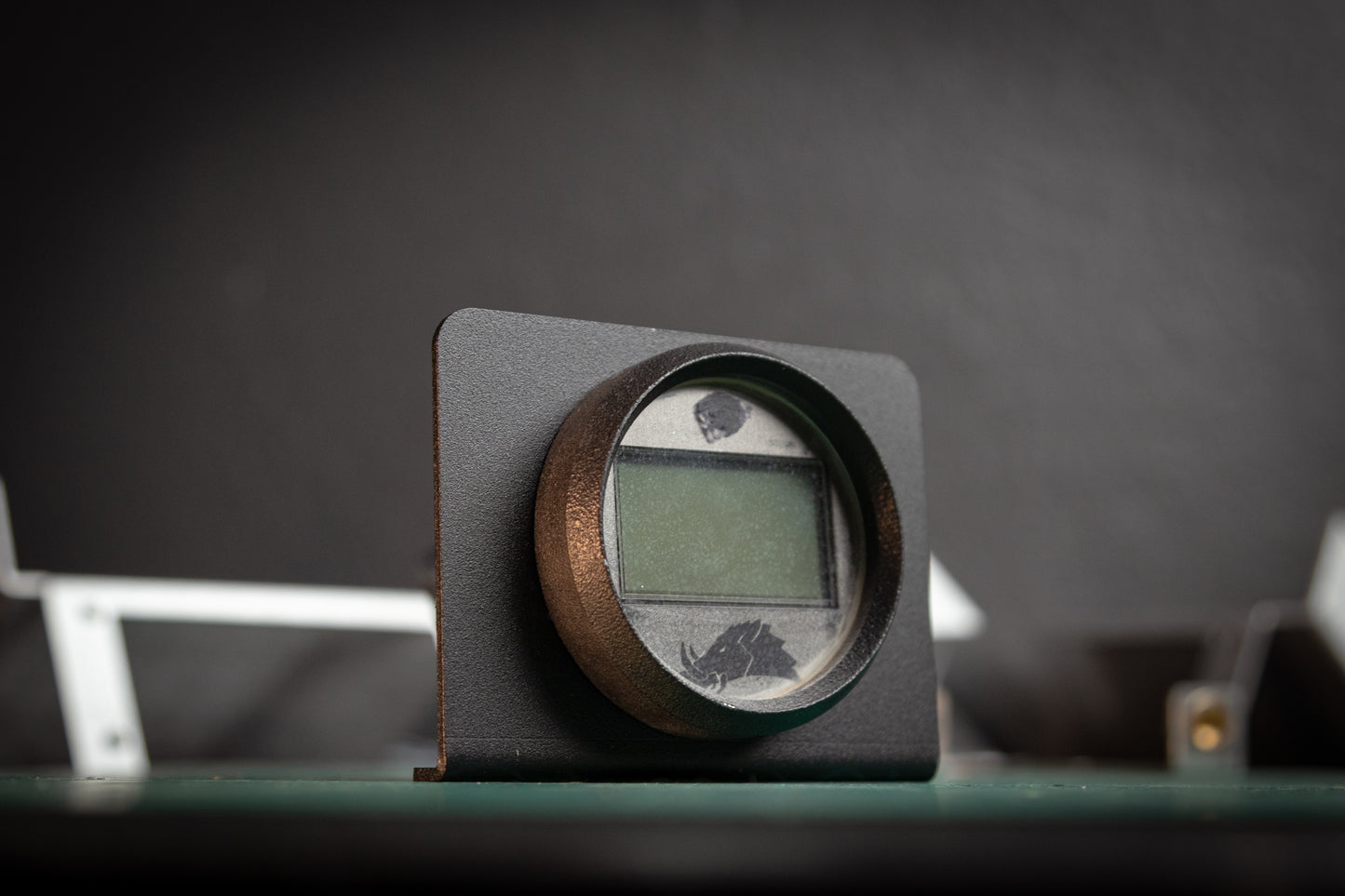 A close-up of the Gauge Mount Sidekick Accessory by StayFlush Motorsports: a small round display in a black frame, mounted on a green circuit board with a blurred background.